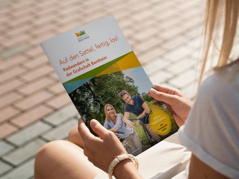 Eine Person mit weißen T-Shirt hält eine Radbroschüre vom Grafschaft Bentheim Tourismus in der Hand